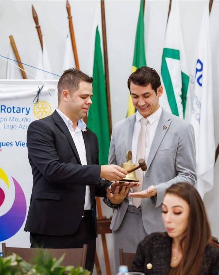 Presidente Marcel (23/24) entregando o sino ao Presidente eleito Eduardo (24/25). O sino representa a disciplina e a ordem que deve dominar o ambiente das reuniões.