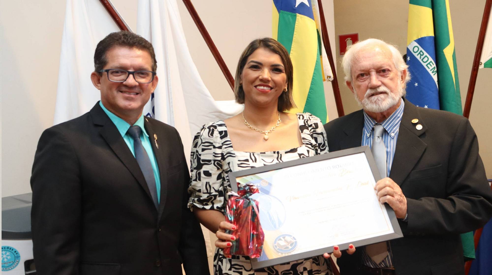 Presidente do RC de Rio Verde 24-25 Rafael de Faria Freire e o Sócio Fundador do RC de Rio Verde Avelar de Moraes Macedo entregam Reconhecimento Rotário á professora premiada.