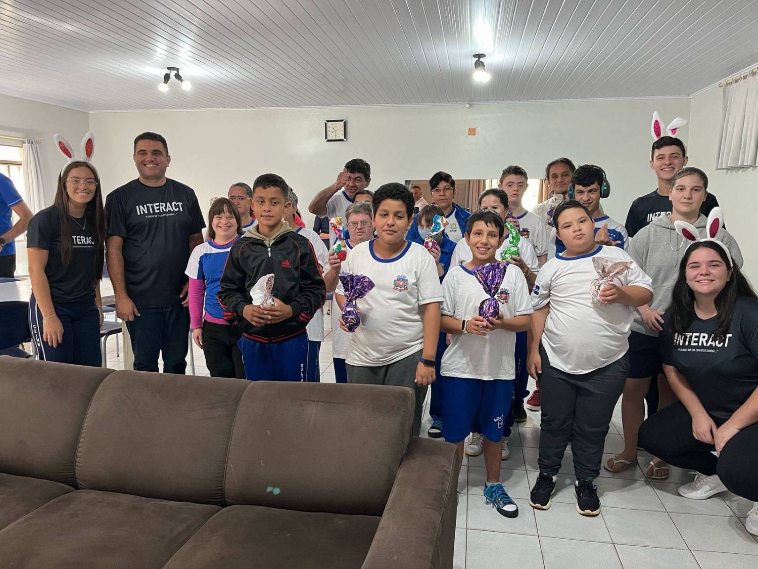 A imagem mostra um grupo de crianças e adultos em um ambiente interno, possivelmente um centro comunitário. Muitas das crianças estão usando uniformes escolares brancos e azuis e todos estão usando tiaras com orelhas de coelho. Eles seguram ovos de chocolate, exibindo sorrisos felizes. Os adultos vestem camisetas pretas com logotipos, incluindo 