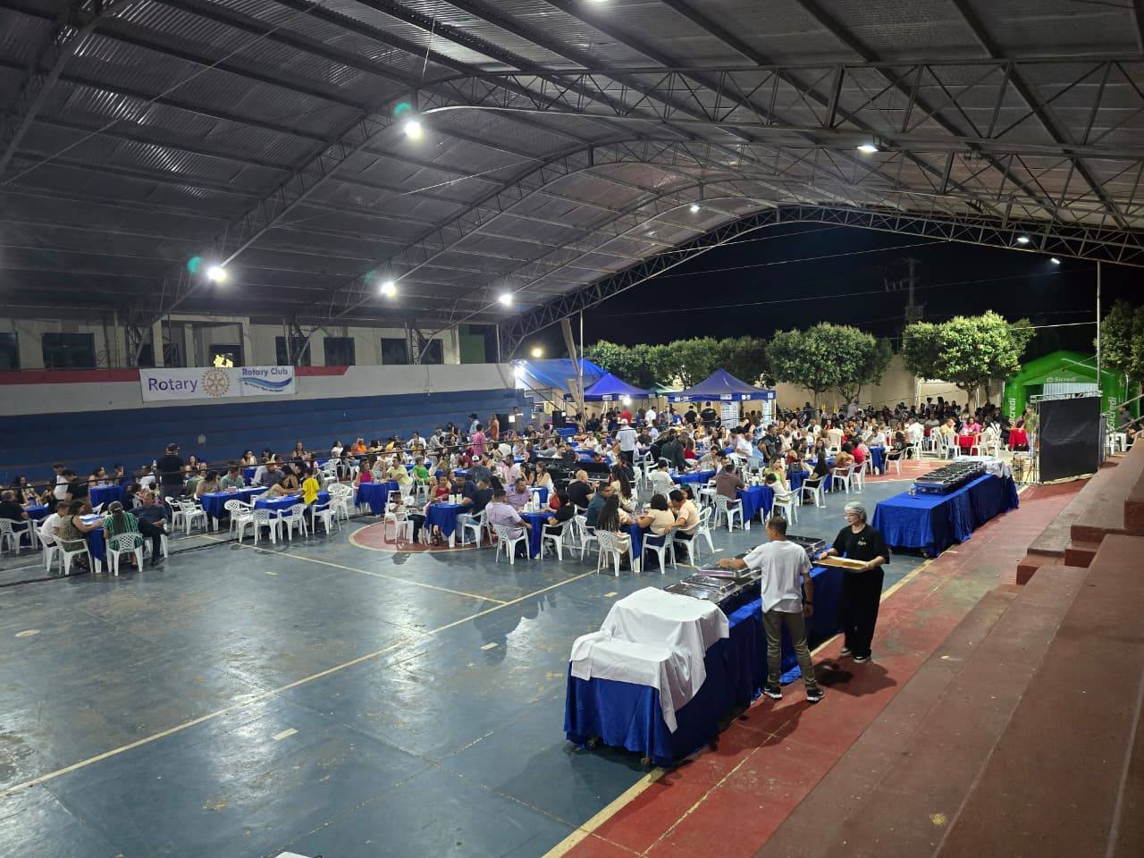 Pessoas sentadas em cadeiras com toalha azul, dentro da quadra da Paroquia São Roque, durante o jantar premiado em Rio Branco.
