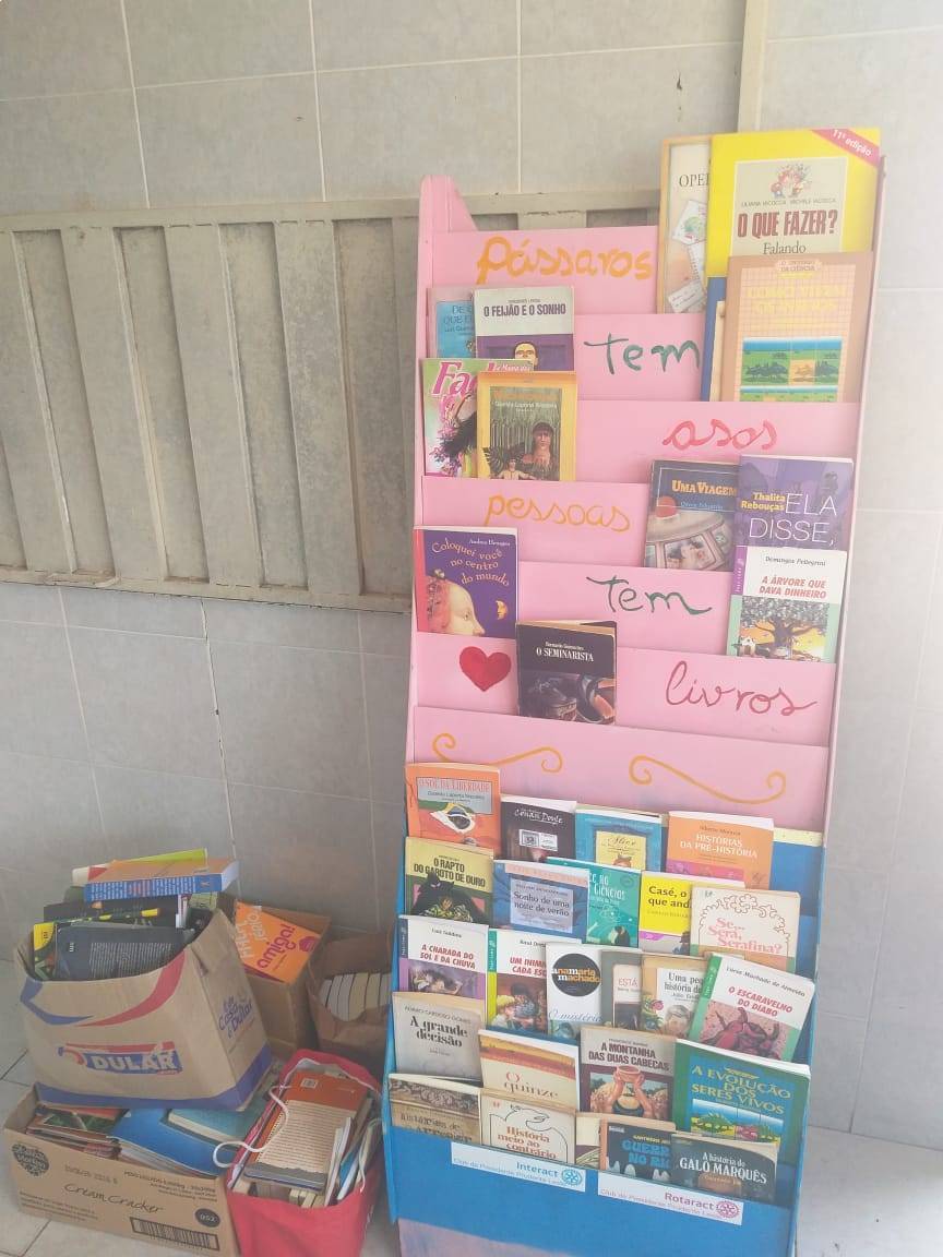 Estante com alguns dos livros, e ao lado da estante, mais livros que foram doados.